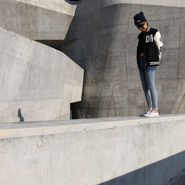 Young woman in varsity jacket on concrete wall, daring to dream.
