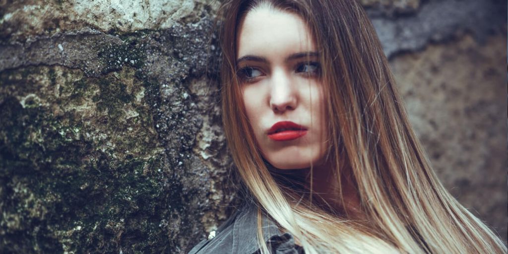 Young woman with long blonde hair, red lipstick, leaning against a mossy wall.