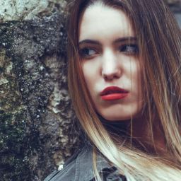 Young woman with long blonde hair, red lipstick, leaning against a mossy wall.