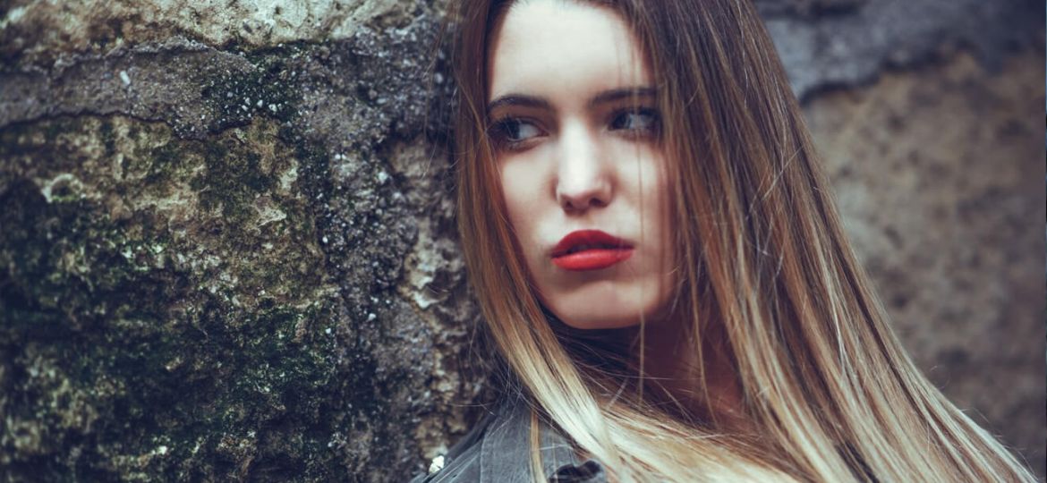 Young woman with long blonde hair, red lipstick, leaning against a mossy wall.