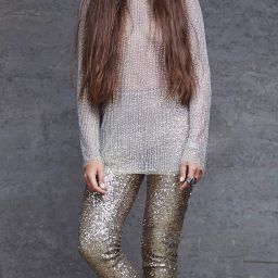 Woman with long brown hair wearing a sparkly silver top and gold sequin pants, posing against a gray wall.
