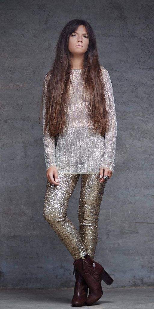 Woman with long brown hair wearing a sparkly silver top and gold sequin pants, posing against a gray wall.