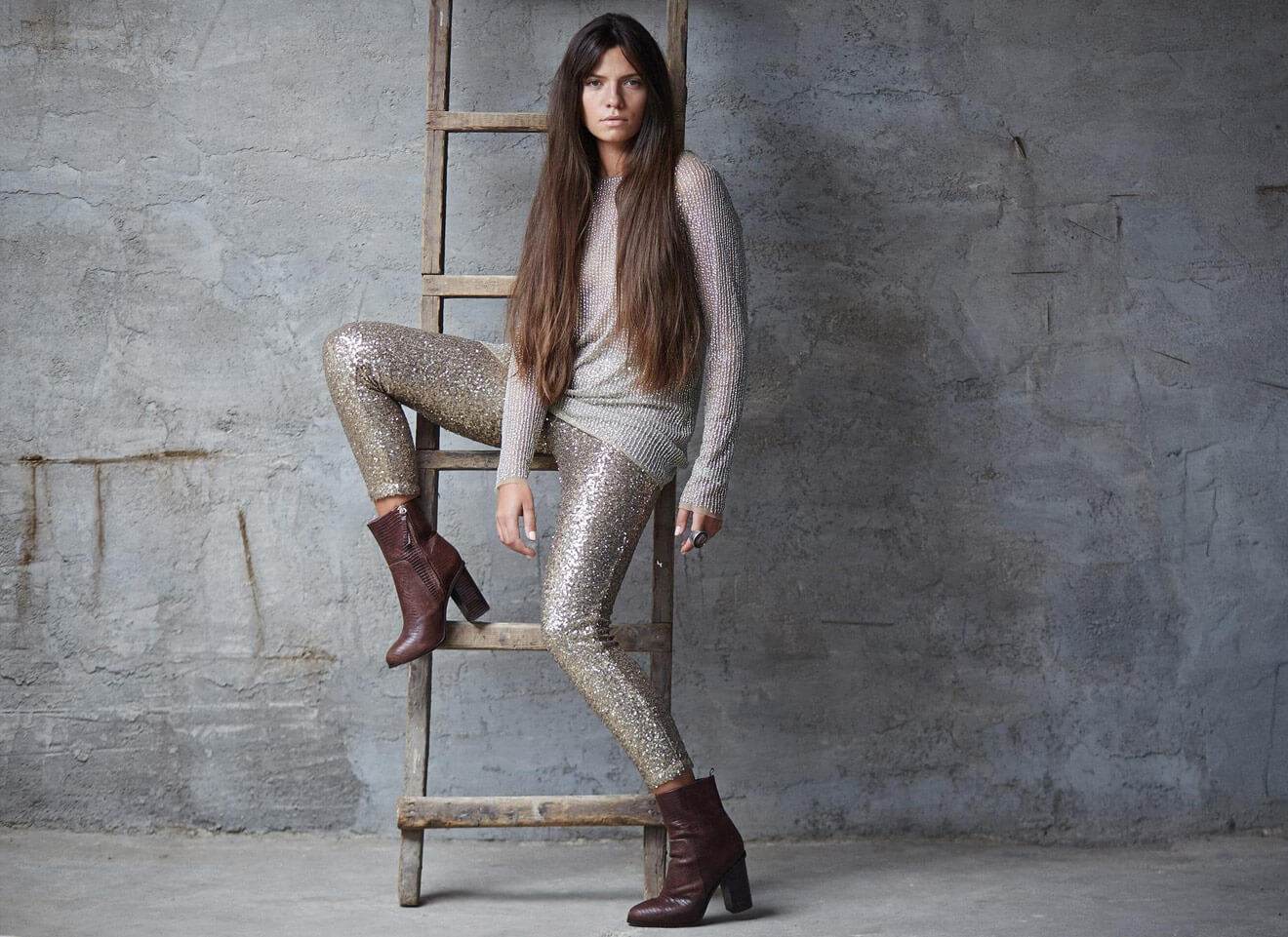 Woman in sparkly leggings and brown boots poses on a wooden ladder against a textured wall.