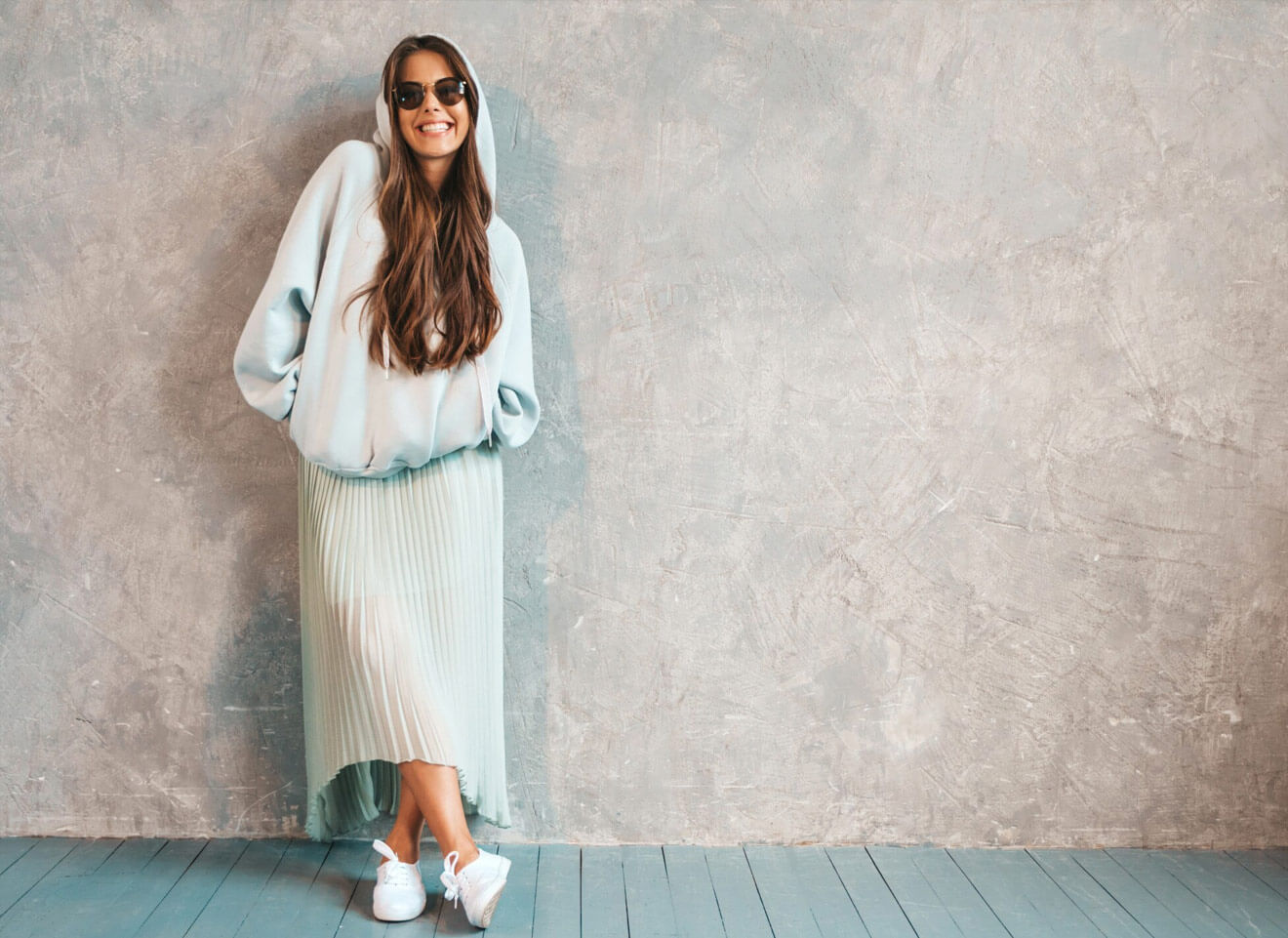 Stylish woman in light blue hoodie and pleated skirt poses against a gray wall.