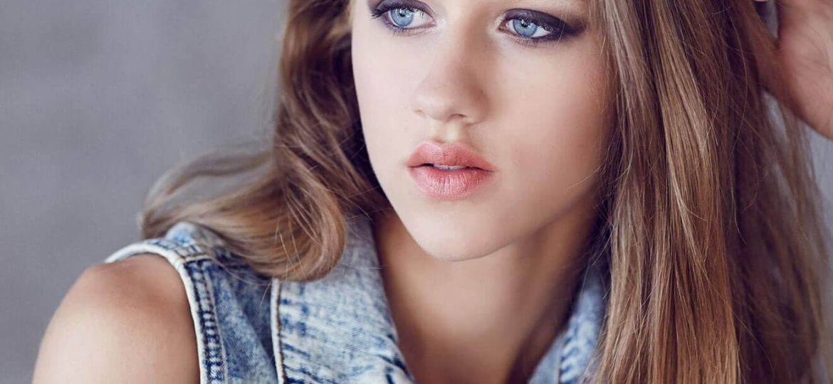 Young woman with long blonde hair, wearing a denim vest.